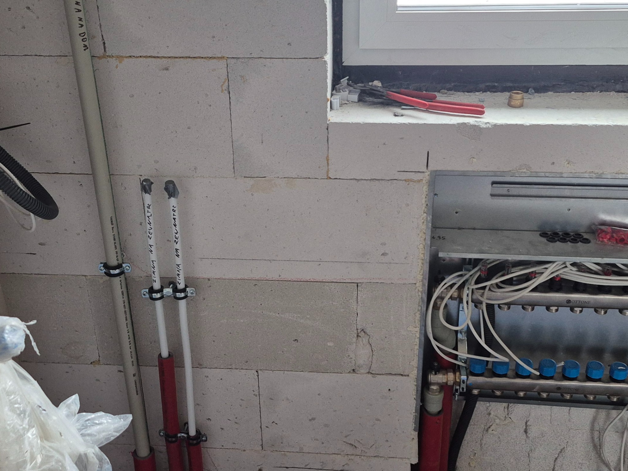 Rozdzielacz ogrzewania podłogowego zamontowany przy ścianie z bloczków, widoczne rury z oznaczeniami, narzędzia leżące na parapecie okna.