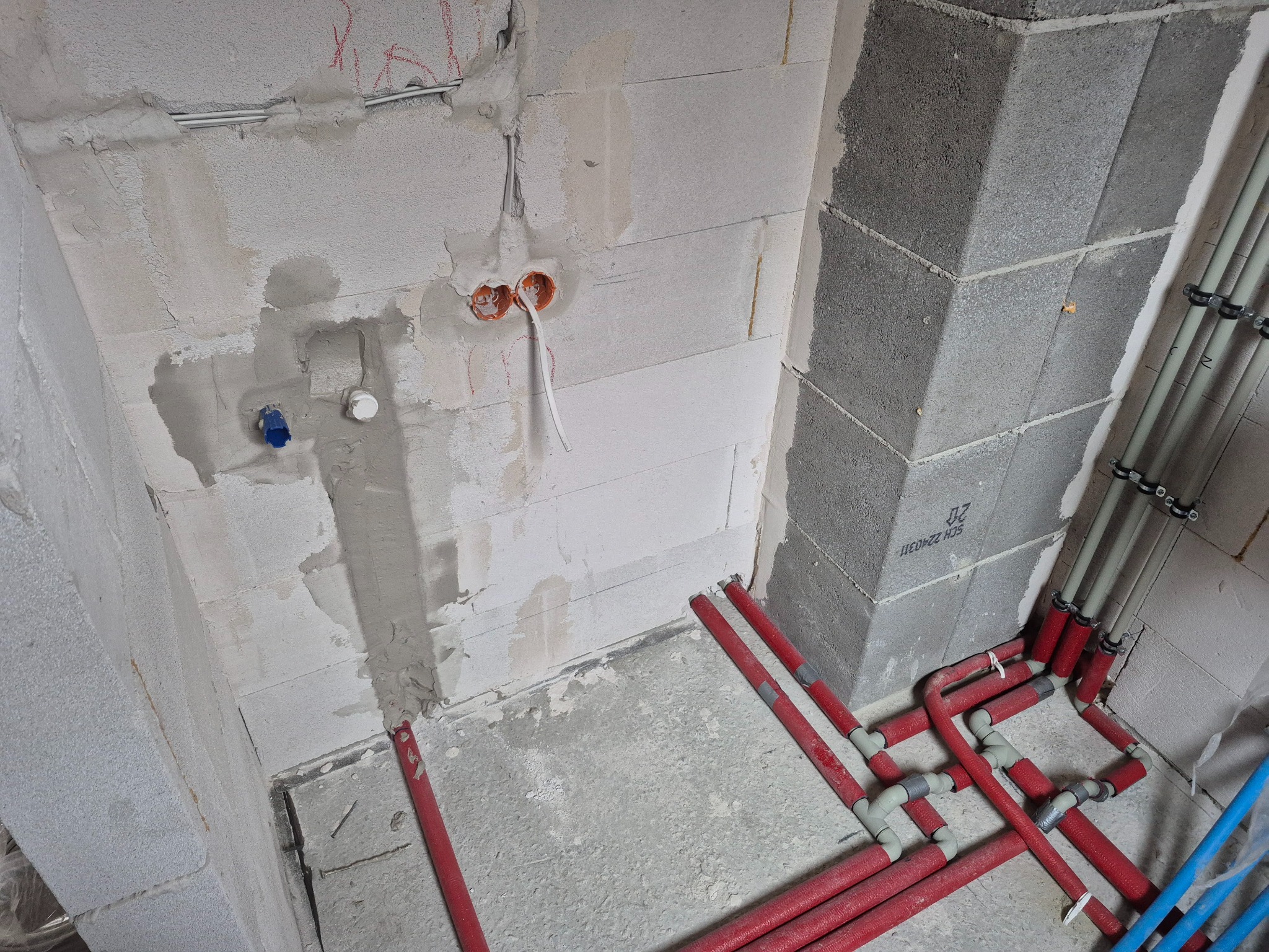 Surowy stan instalacji w nowym budynku: czerwone rury grzewcze w izolacji, szare rury wodociągowe mocowane do ściany z betonu komórkowego, puszki elektryczne w ścianie, przygotowane do podłączenia.