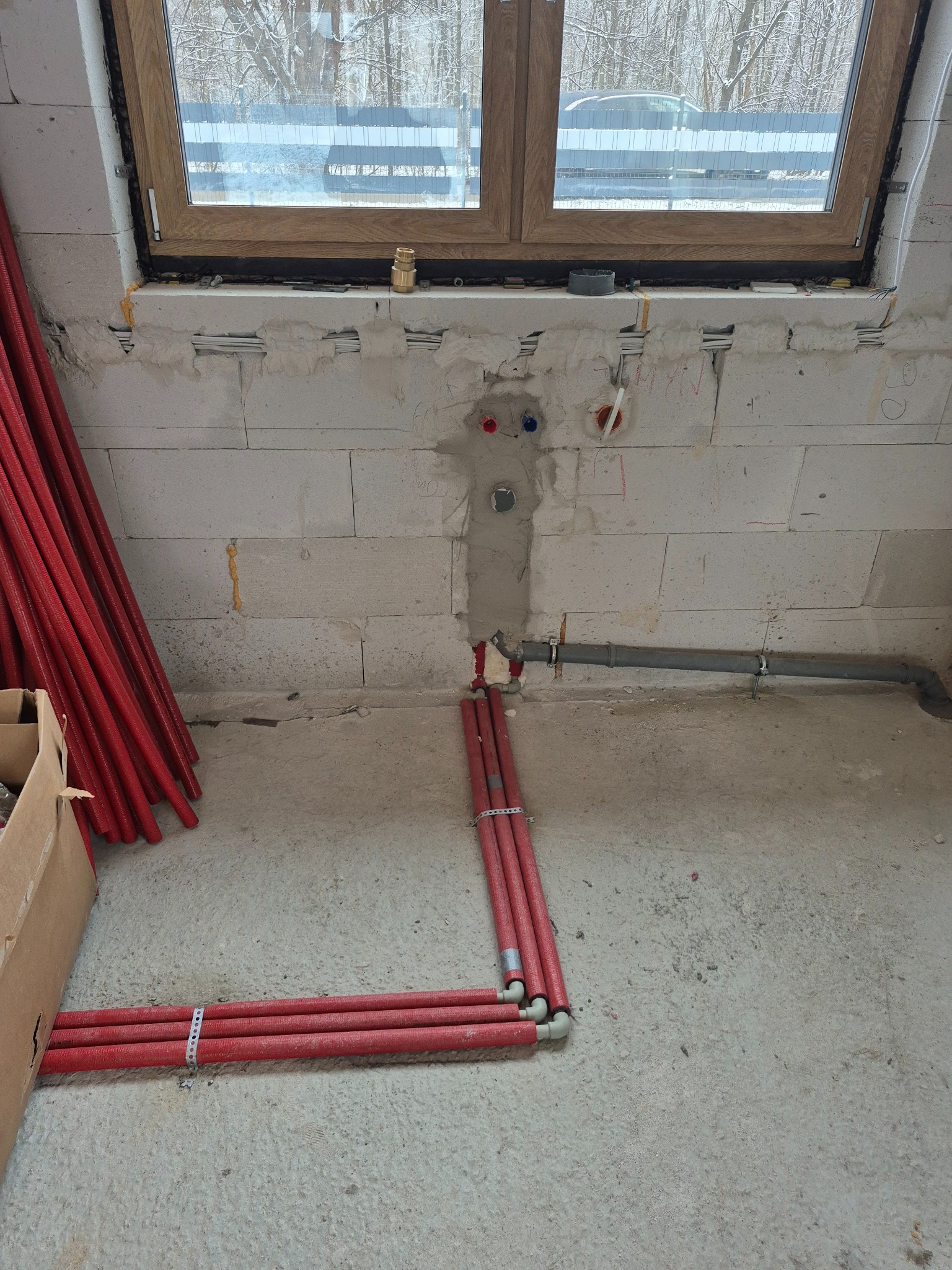 Czerwone rury w izolacji ułożone na podłodze w surowym stanie, z widocznymi przyłączami i fragmentem szarej rury kanalizacyjnej, na tle okna z widokiem na zimowy krajobraz.