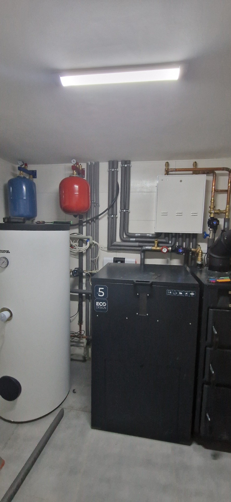 Kotłownia z piecem na paliwo stałe, zbiornikiem akumulacyjnym i instalacją hydrauliczną z rurami w izolacji, widoczne naczynia wzbiorcze w kolorze niebieskim i czerwonym.