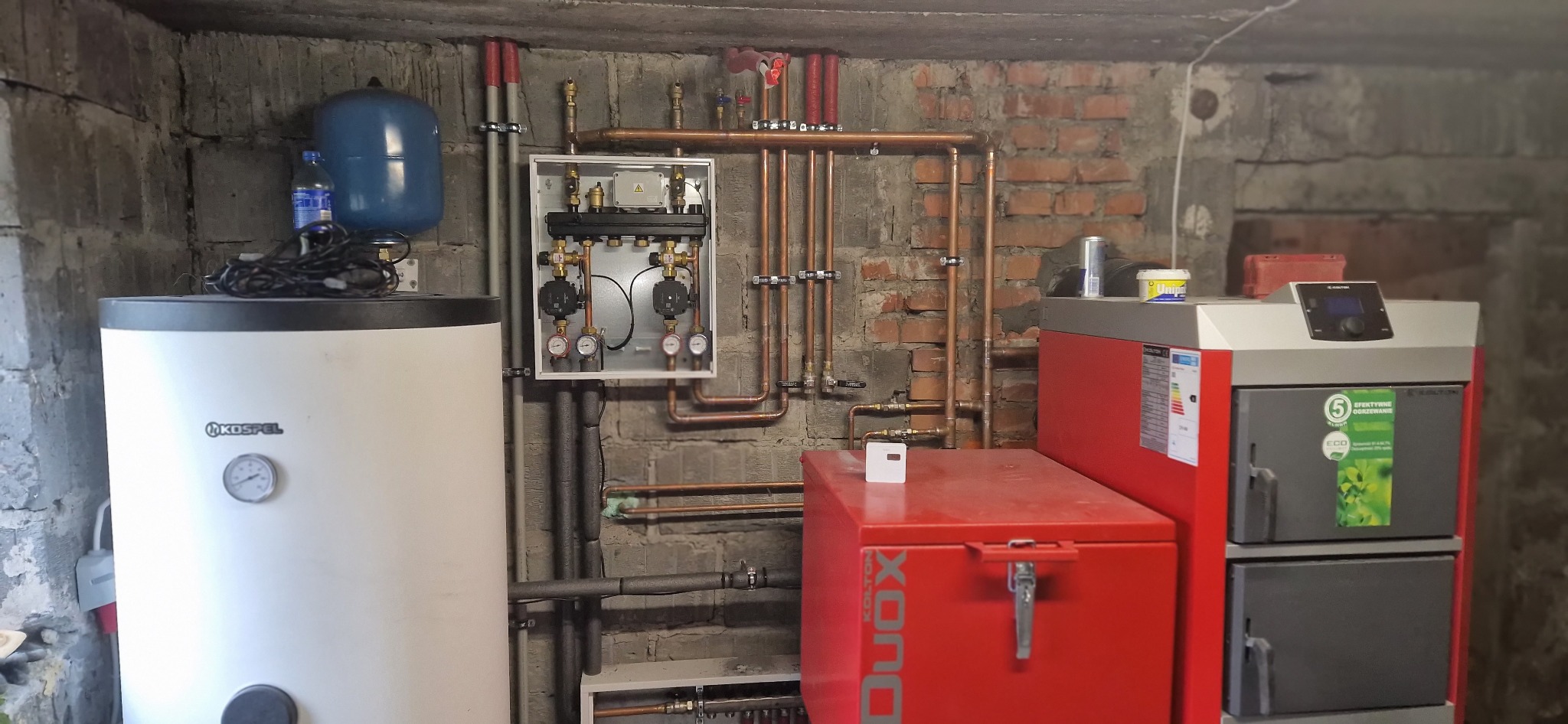 Instalacja grzewcza w kotłowni: biały zbiornik Kospel, czerwony zasobnik Kolton Duox, rozdzielacz w obudowie, miedziane rury, manometry i zawory na tle surowej ściany z cegły i betonu, butelka wody...