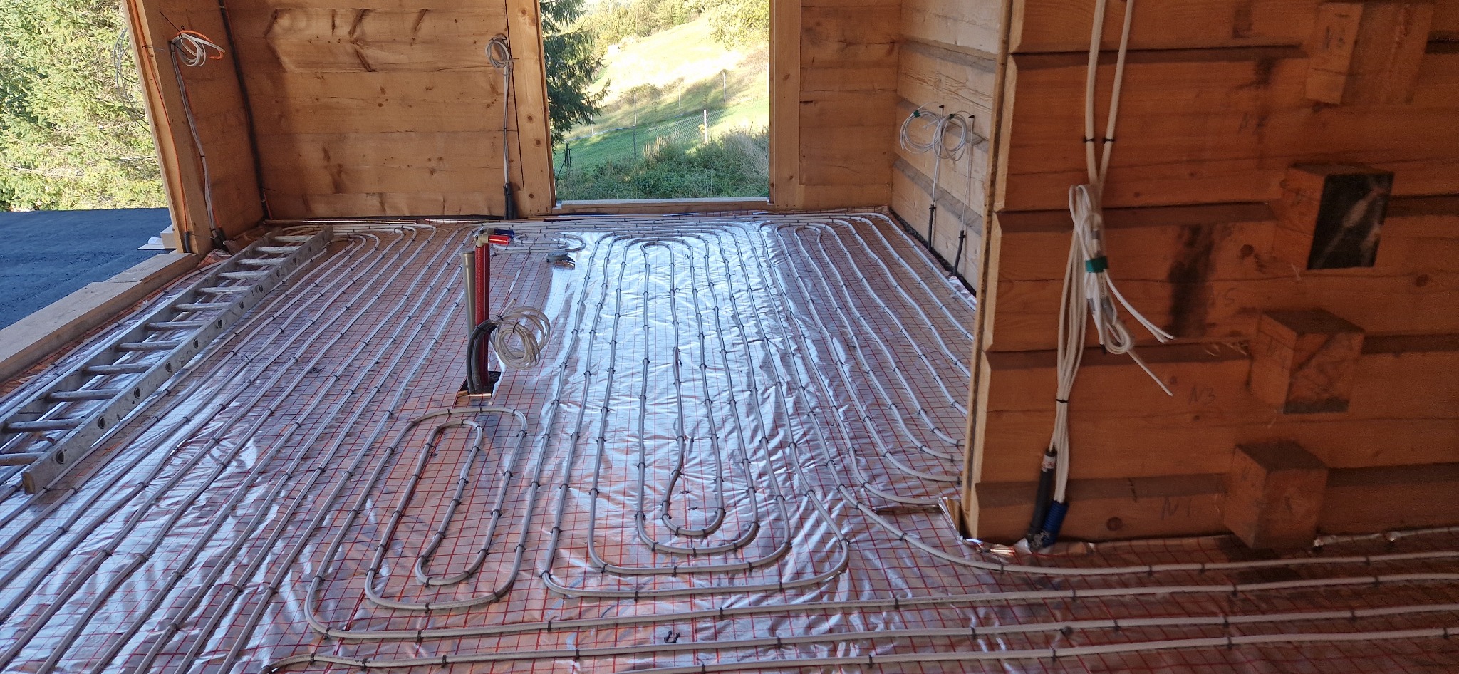 Montaż rur grzewczych podłogowych na folii odblaskowej z siatką w drewnianym domu w trakcie budowy, widoczne przewody i drabina.