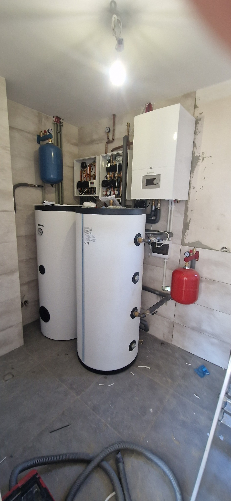 Dwa białe zasobniki ciepłej wody w kotłowni, z widoczną instalacją hydrauliczną, piecem gazowym na ścianie i otwartą skrzynką z zaworami.