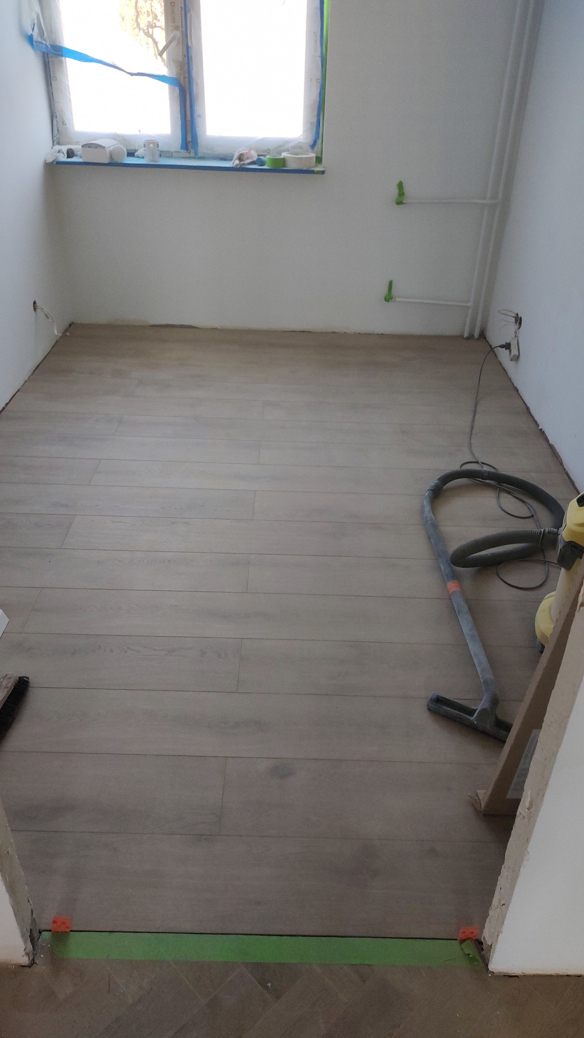 Ułożona podłoga z paneli imitujących drewno w jasnym odcieniu, widoczny odkurzacz przemysłowy, okno zabezpieczone folią malarską oraz listwa przypodłogowa w kolorze zielonym.