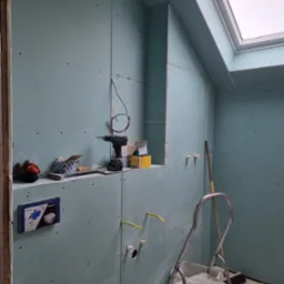 Łazienka w trakcie remontu: ściany wyłożone płytami kartonowo-gipsowymi, widoczne instalacje hydrauliczne, narzędzia (wiertarka, młotek) na półce, drabina i okno dachowe.