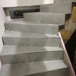 Klatka schodowa w trakcie wykańczania z szarymi płytkami, widoczne krzyżyki dystansowe, fragment okna i ściana w stanie surowym.