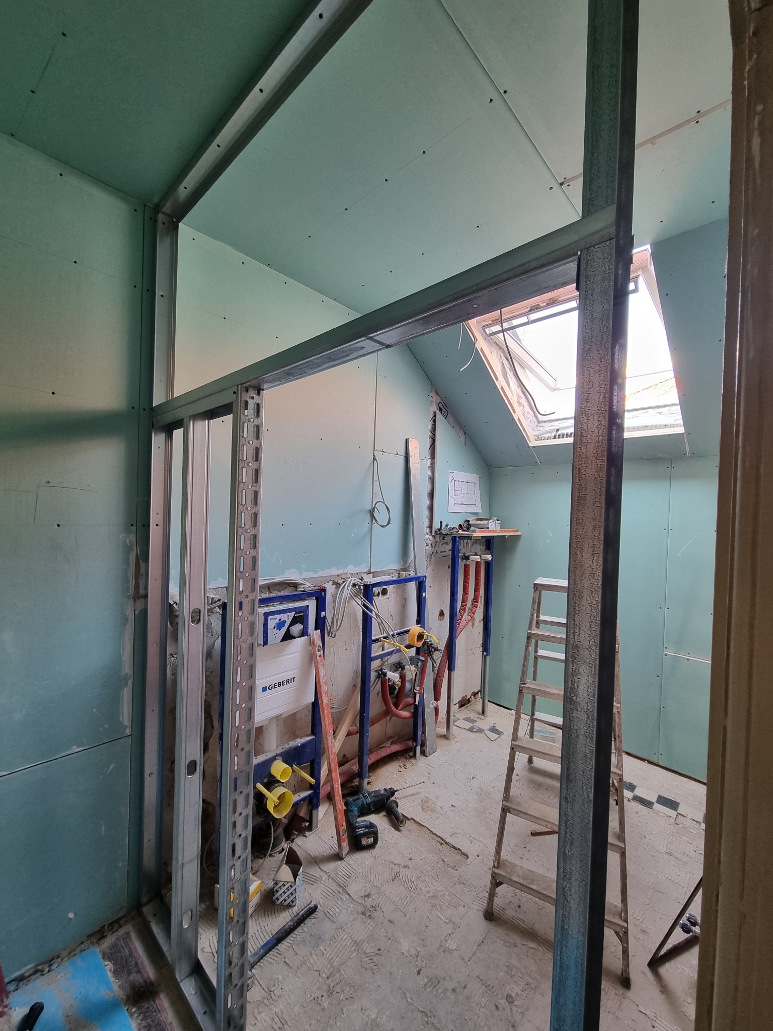 Łazienka w trakcie remontu: widoczne stelaże podtynkowe Geberit, konstrukcja z profili metalowych, płyty gipsowo-kartonowe na ścianach, drabina i narzędzia na podłodze, okno dachowe w tle.