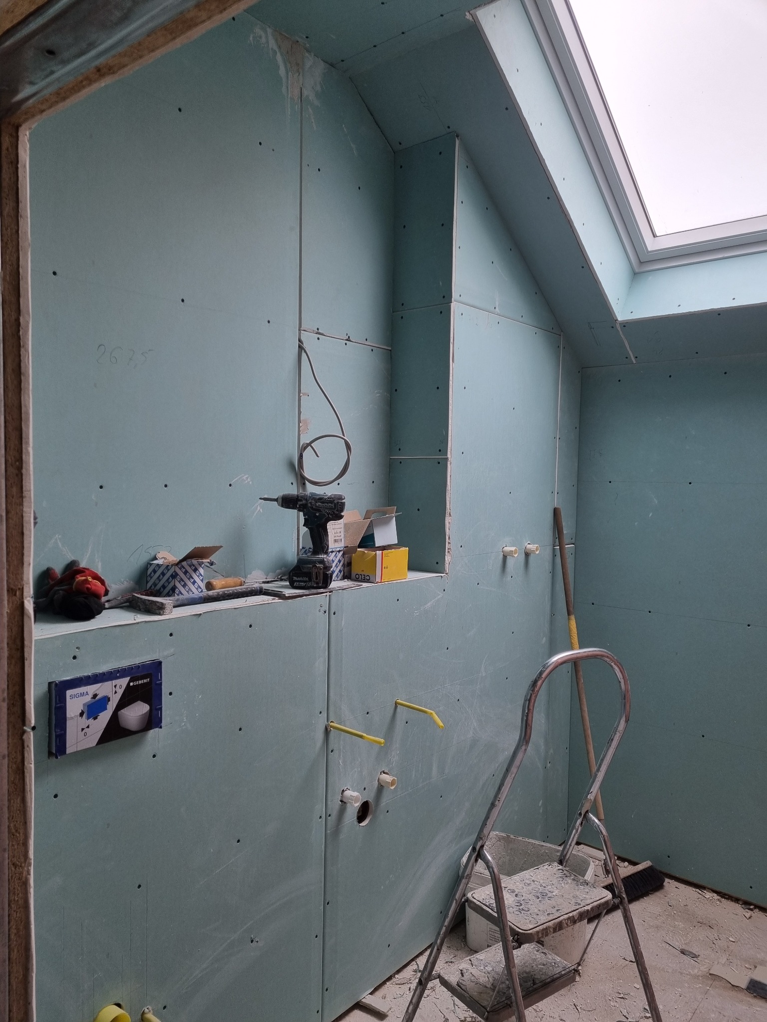 Łazienka w trakcie remontu: ściany wyłożone płytami kartonowo-gipsowymi, widoczne instalacje hydrauliczne, narzędzia (wiertarka, młotek) na półce, drabina i okno dachowe.
