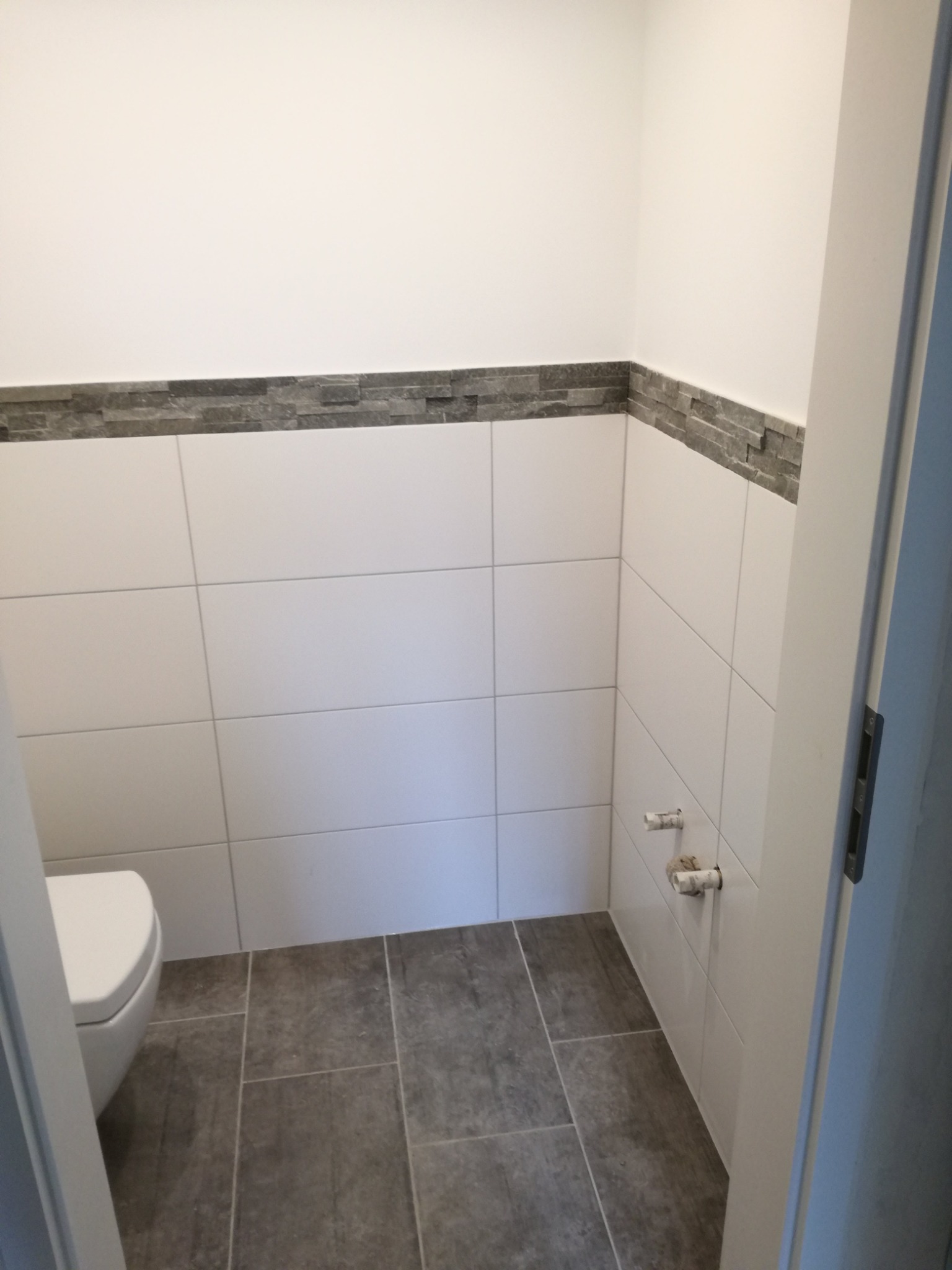 Nowoczesna łazienka z białymi płytkami ściennymi, szarą dekoracyjną listwą i podłogą z szarych płytek, widoczne przyłącza wodne i częściowo toaleta.