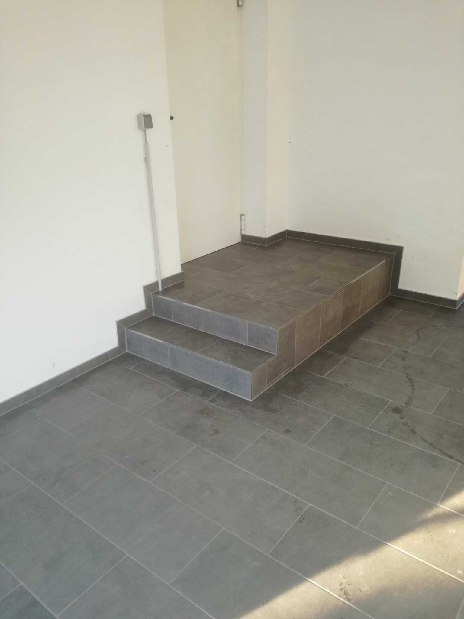 Dwu-stopniowe schody wykończone szarą, prostokątną płytką ceramiczną, dopasowaną do podłogi, z widoczną listwą przypodłogową i gładką, białą ścianą w tle.