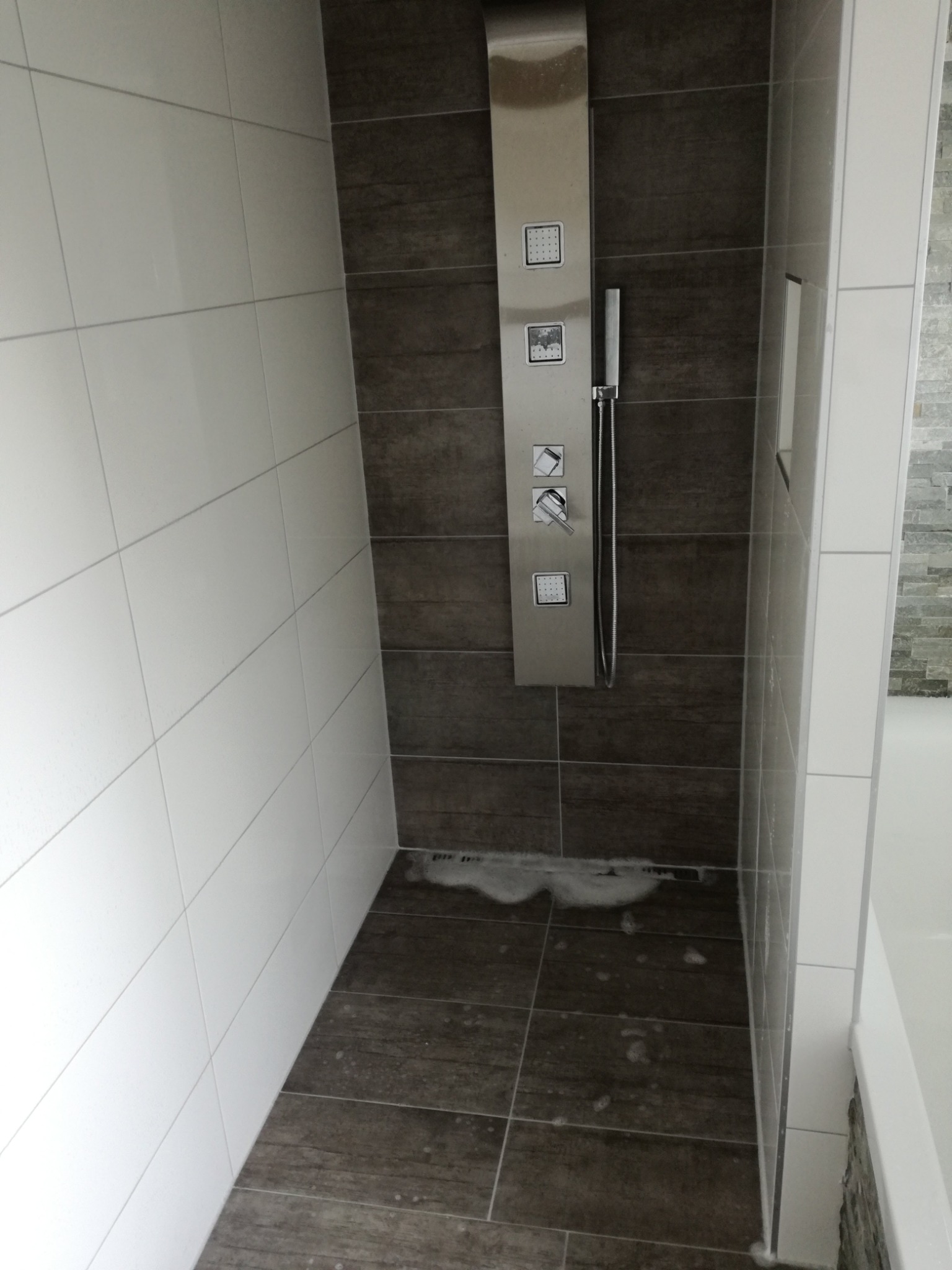 Nowoczesna kabina prysznicowa z panelem natryskowym ze stali nierdzewnej, wykończona białymi i brązowymi płytkami ceramicznymi z widocznymi śladami wody po użyciu.