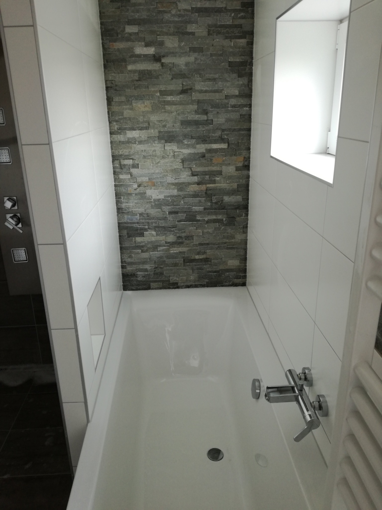 Wanna w nowoczesnej łazience z szarą ścianą wyłożoną kamieniem dekoracyjnym, białe płytki na pozostałych ścianach, minimalistyczna bateria wannowa.