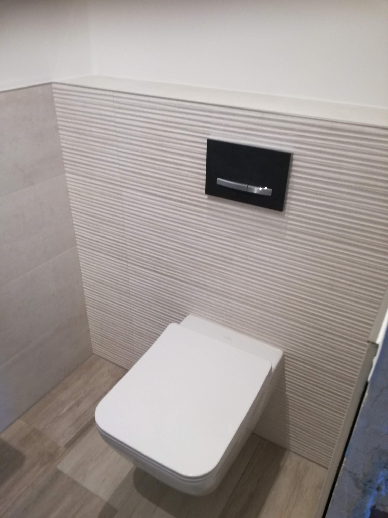 Nowoczesna toaleta podwieszana z czarnym panelem spłukującym na ścianie wykończonej płytkami z poziomymi żłobieniami, na podłodze imitacja drewna.