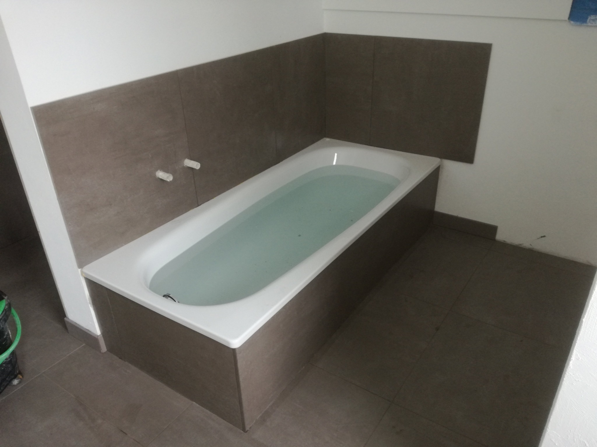 Wanna wypełniona wodą, obudowana płytkami w odcieniach szarości, z fragmentem wykończonej ściany w podobnych płytkach i widocznym fragmentem podłogi z płytek.