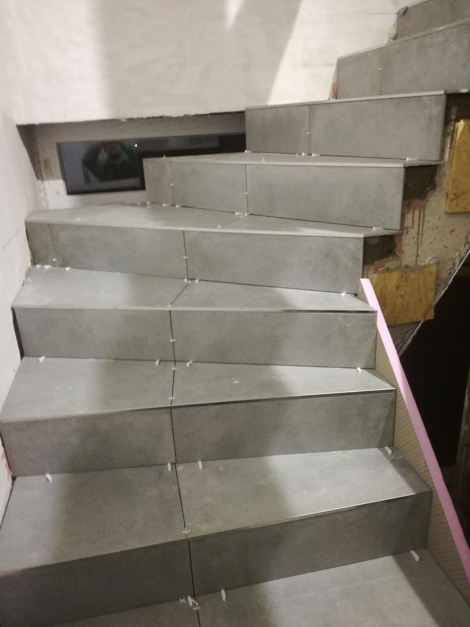 Klatka schodowa w trakcie wykańczania z szarymi płytkami, widoczne krzyżyki dystansowe, fragment okna i ściana w stanie surowym.