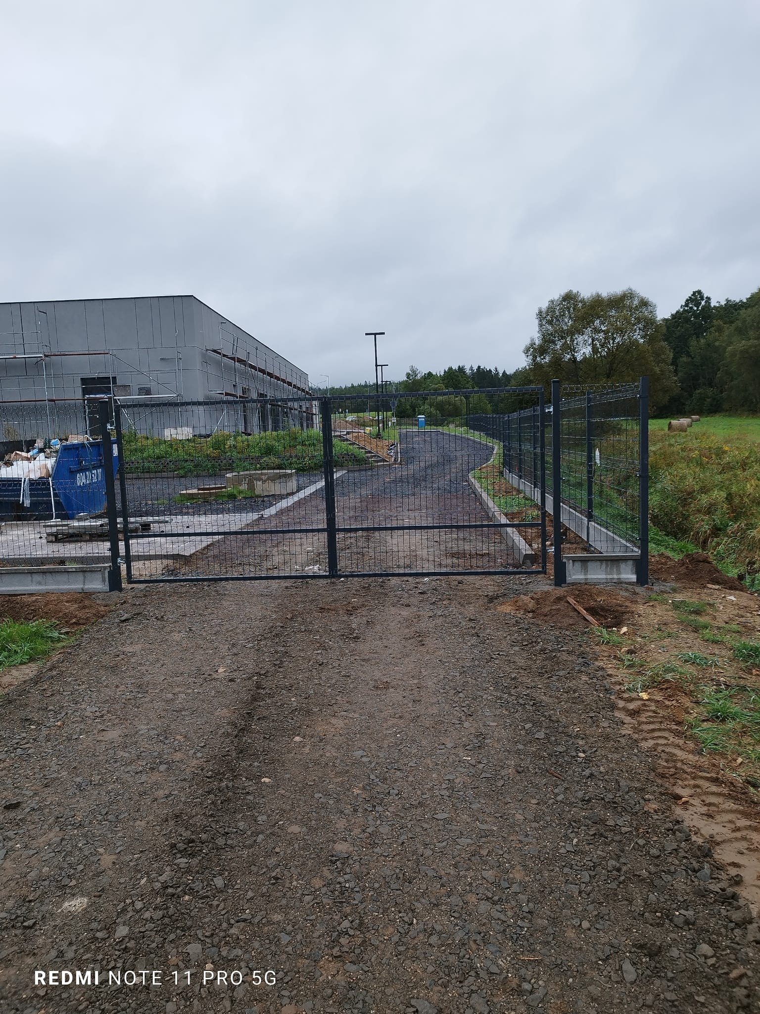 Brama z siatki z czarnymi słupkami na wjeździe do posesji, widok z zewnątrz, w tle budynek w trakcie budowy i zieleń.