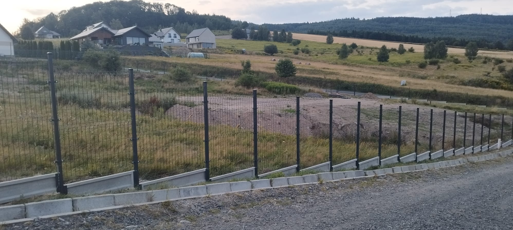 Długi, prosty odcinek ogrodzenia panelowego z podmurówką betonową, na nierównym terenie z widokiem na domy i pola.