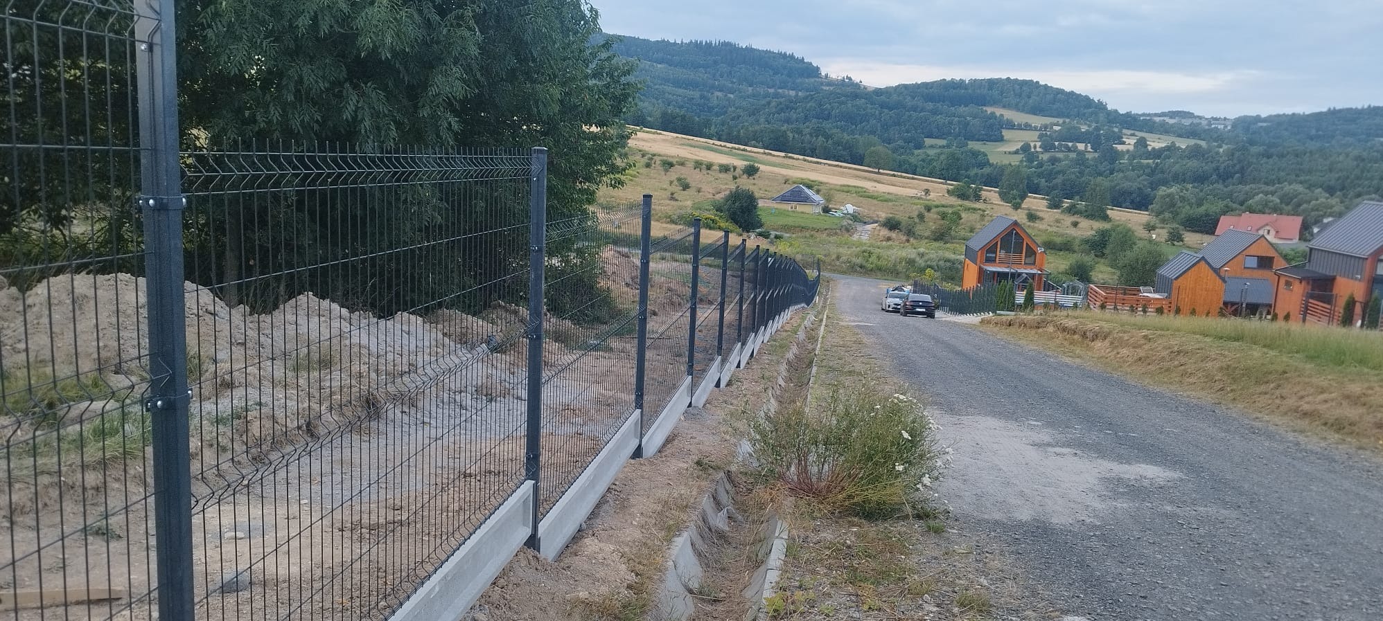 Długa linia czarnego ogrodzenia panelowego z betonową podmurówką, ciągnąca się wzdłuż polnej drogi na tle pagórkowatego krajobrazu z domami.