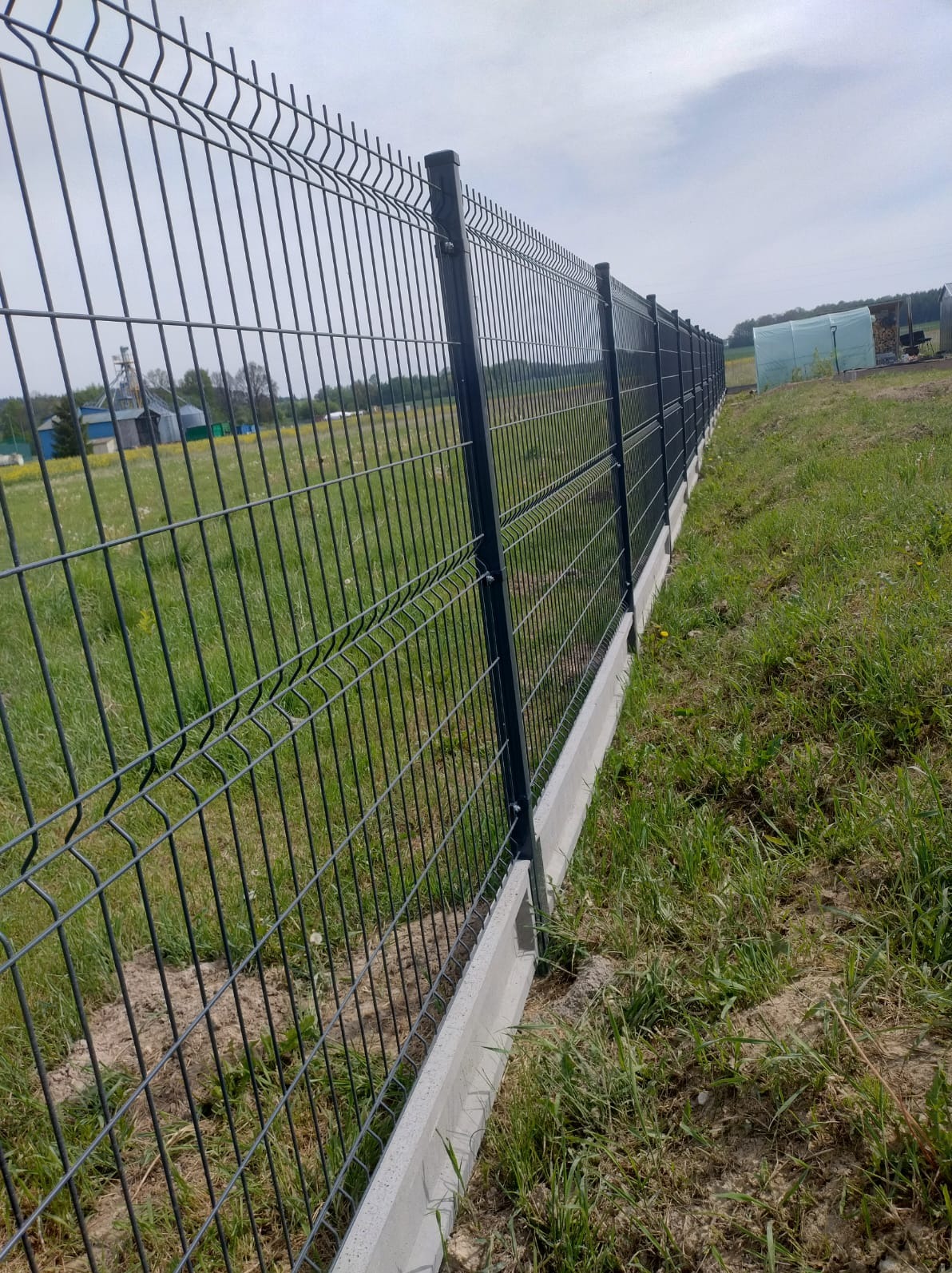 Długi, ciemny system ogrodzeniowy z paneli z betonową podmurówką, widziany z boku na tle zielonej łąki i zabudowań gospodarczych.
