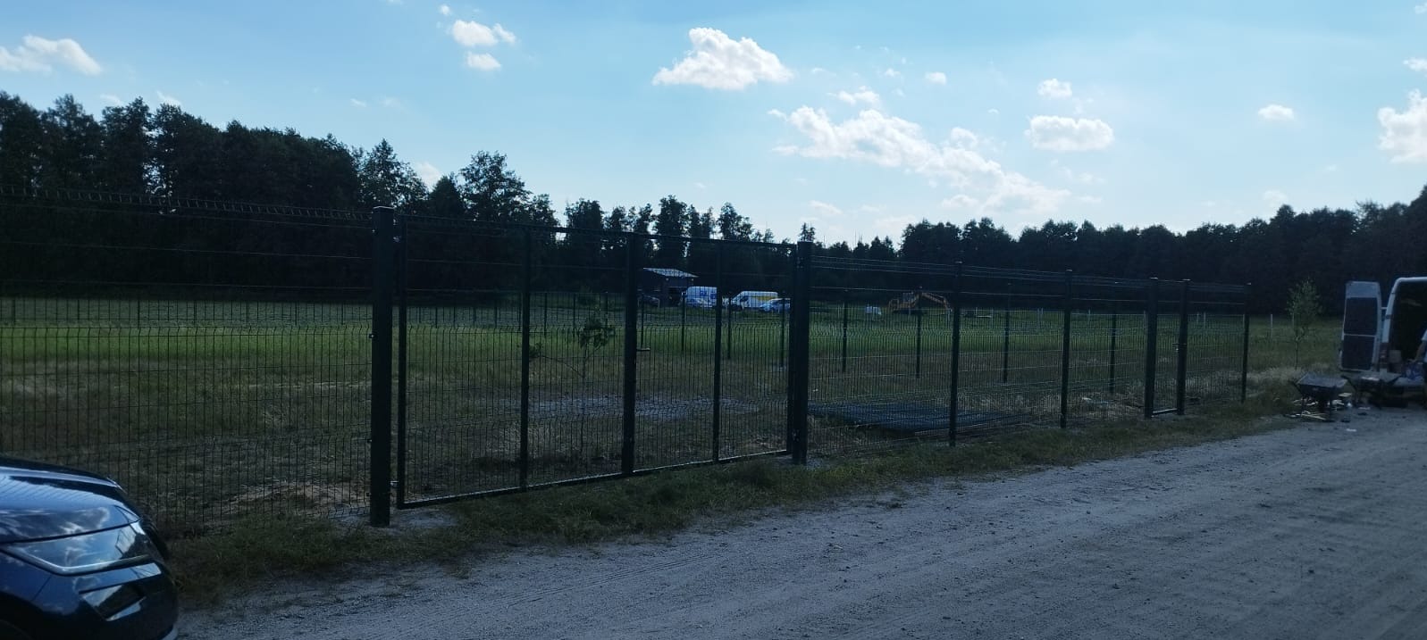 Ciemne panele ogrodzeniowe z bramą wjazdową, widoczne na tle zielonego pola i lasu, z fragmentem samochodu i furgonetki w tle, sugerujące trwający montaż.