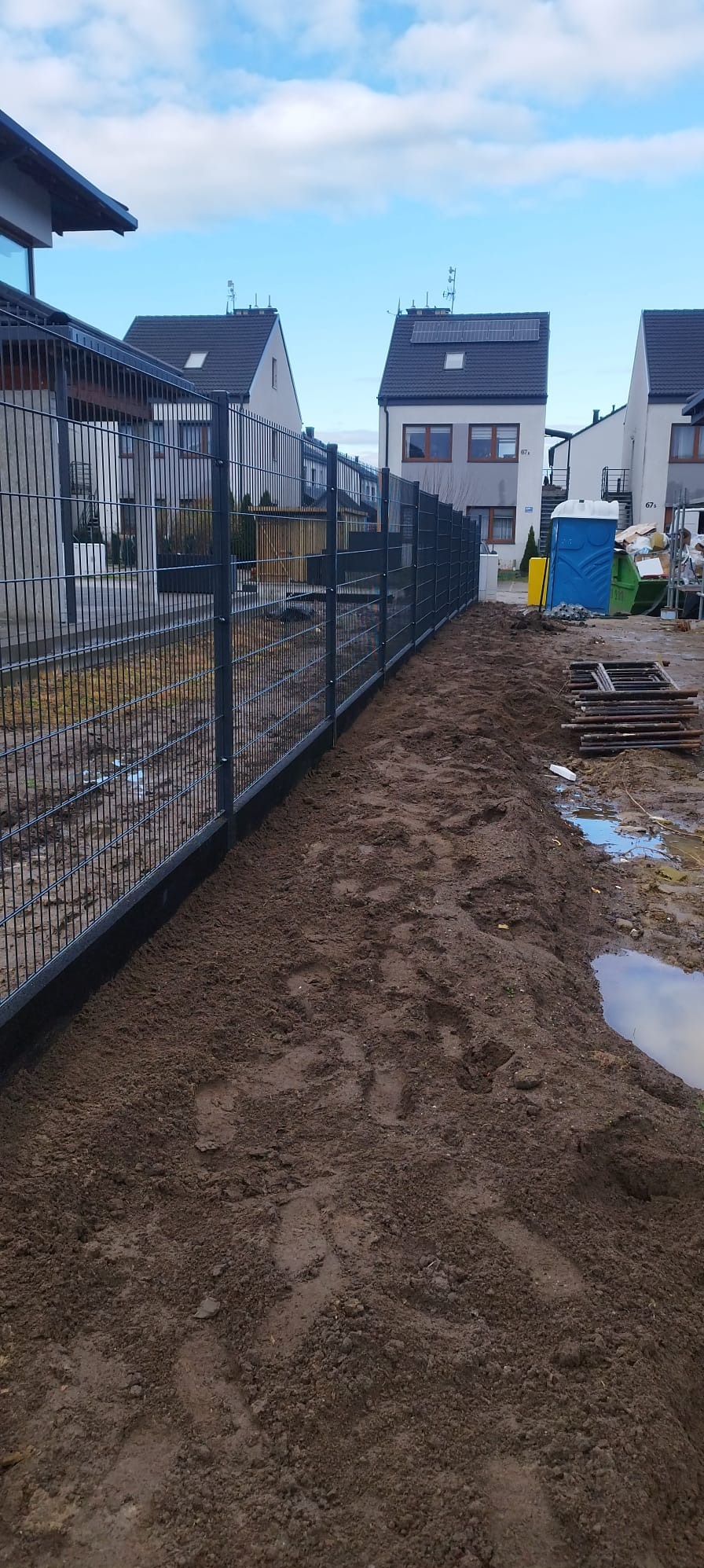 Szary system ogrodzeniowy z paneli na posesji z widocznymi domami w tle, teren wokół ogrodzenia przygotowany pod nasadzenia, widoczna przenośna toaleta i resztki materiałów budowlanych.
