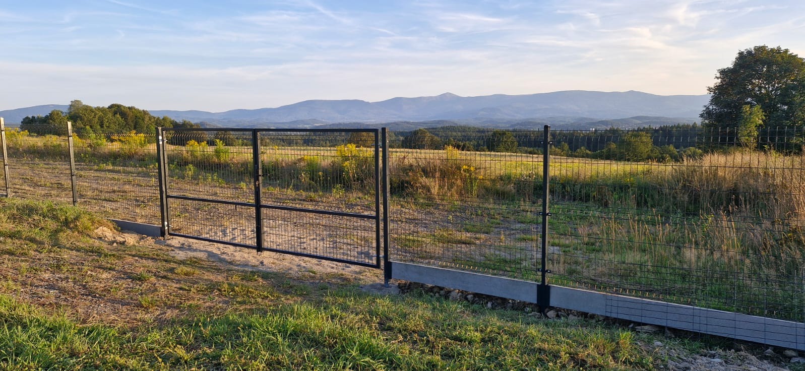 Czarna brama z siatki na betonowej podmurówce, z widokiem na pagórkowaty krajobraz z lasami i górami w tle, wykonana w słoneczny dzień.