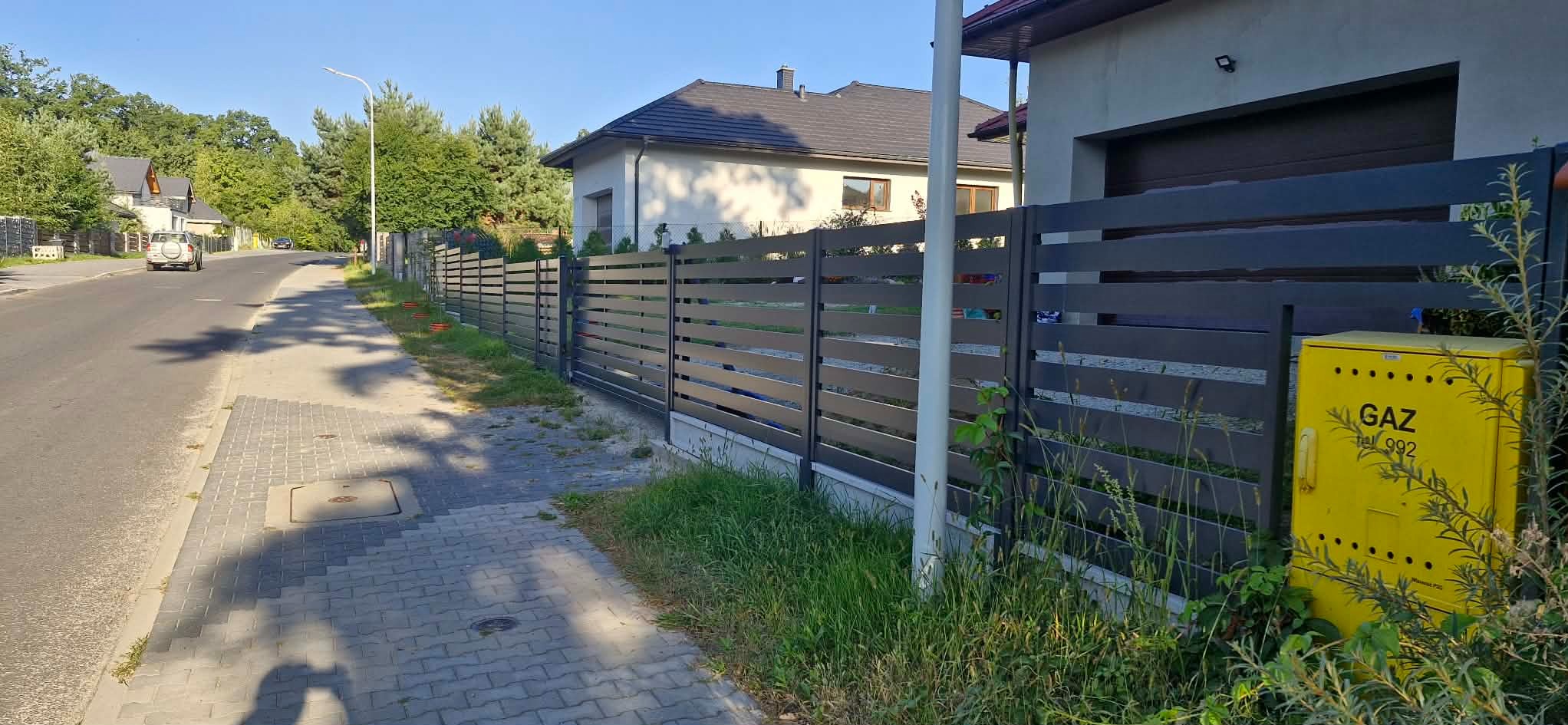 Nowoczesne ogrodzenie posesji z poziomych paneli w Mienicach. Szara kolorystyka, widok z chodnika, skrzynka gazowa przy ogrodzeniu. Estetyczne wykończenie.