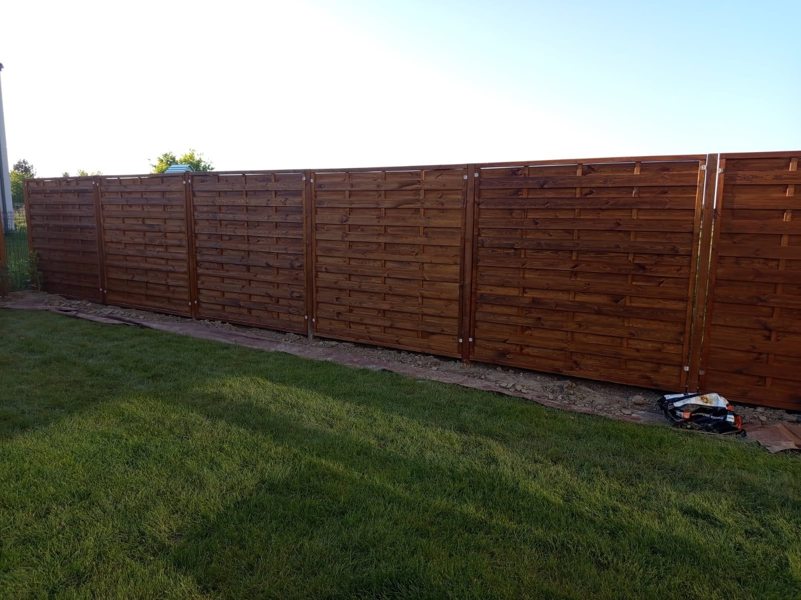 Ciemnobrązowe, poziome panele ogrodzeniowe z drewna, oddzielone metalowymi słupkami, na tle zielonego trawnika i błękitnego nieba. Widoczne narzędzia montażowe.