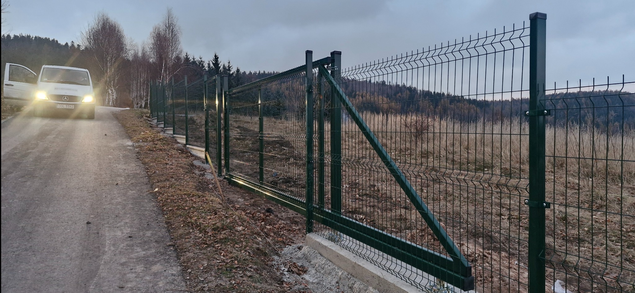Nowoczesne, zielone ogrodzenie panelowe z bramą, zamontowane na betonowej podmurówce. W tle zalesione wzgórza i biały bus z otwartymi drzwiami.