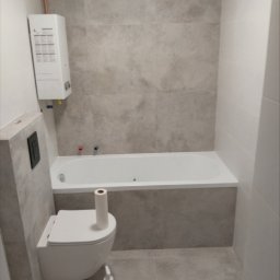 A.D.G - Nowoczesna łazienka z wanną, podwieszanym WC, grzejnikiem przepływowym i szarą płytką imitującą kamień na ścianach i podłodze. Na podłodze stoi butelka środka czyszczącego i rolka papieru...