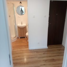 A.D.G - Ukończona łazienka z kabiną prysznicową, umywalką z szafką i toaletą, widok z korytarza na drewnopodobną podłogę.