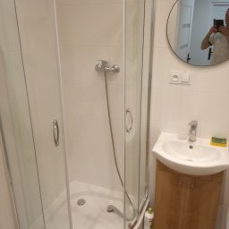 A.D.G - Nowoczesna łazienka po remoncie: kabina prysznicowa z chromowaną armaturą, okrągłe lustro z odbiciem osoby robiącej zdjęcie telefonem, umywalka z szafką w kolorze drewna i brązowe płytki podłogowe.