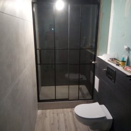 A.D.G - Niedokończona łazienka z płytkami w odcieniach szarości, kabiną prysznicową z czarnymi ramami i podwieszaną toaletą; widoczne narzędzia i materiały budowlane na blacie.