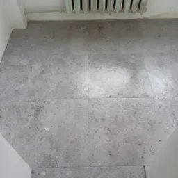 Szare, matowe płytki podłogowe ułożone w kwadraty, widoczne fugi, przy ścianie stary żeliwny grzejnik pomalowany na zielono, fragment futryny drzwiowej.