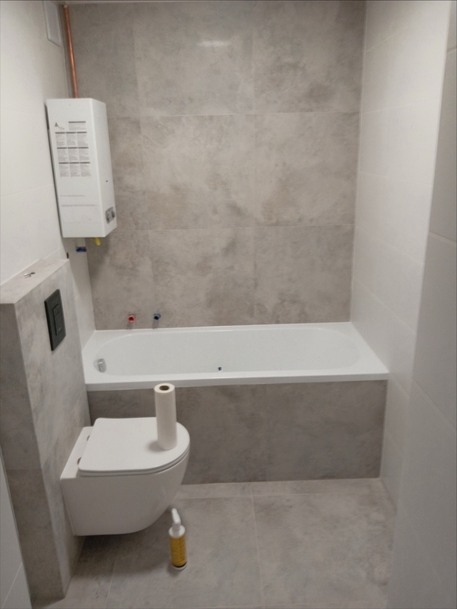 Nowoczesna łazienka z wanną, podwieszanym WC, grzejnikiem przepływowym i szarą płytką imitującą kamień na ścianach i podłodze. Na podłodze stoi butelka środka czyszczącego i rolka papieru...