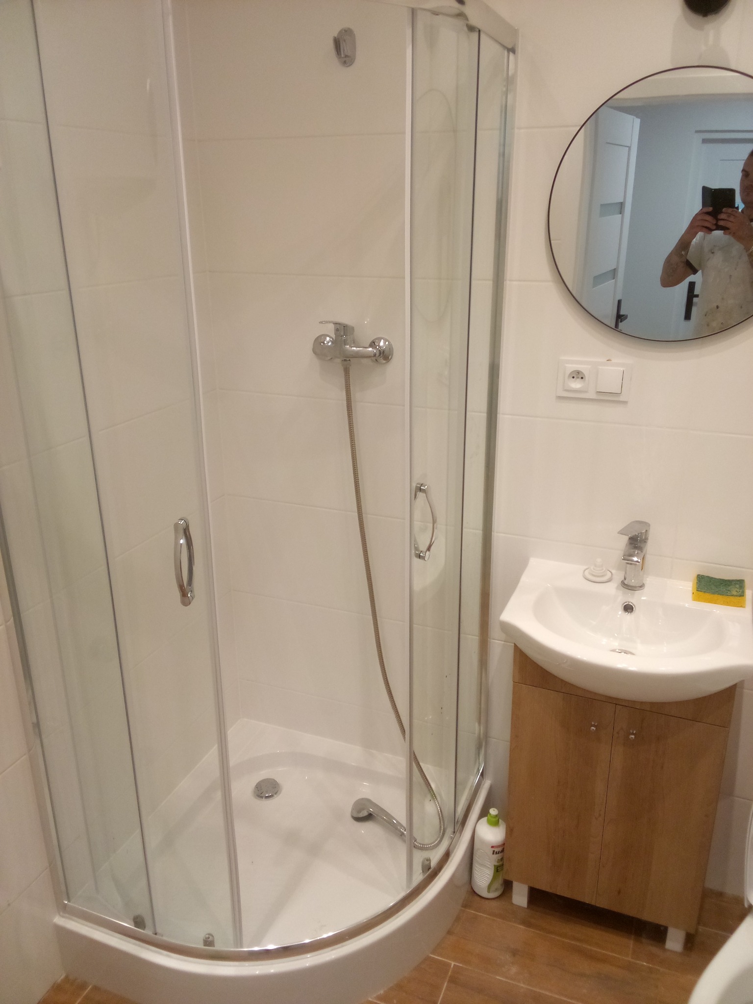 Nowoczesna łazienka po remoncie: kabina prysznicowa z chromowaną armaturą, okrągłe lustro z odbiciem osoby robiącej zdjęcie telefonem, umywalka z szafką w kolorze drewna i brązowe płytki podłogowe.