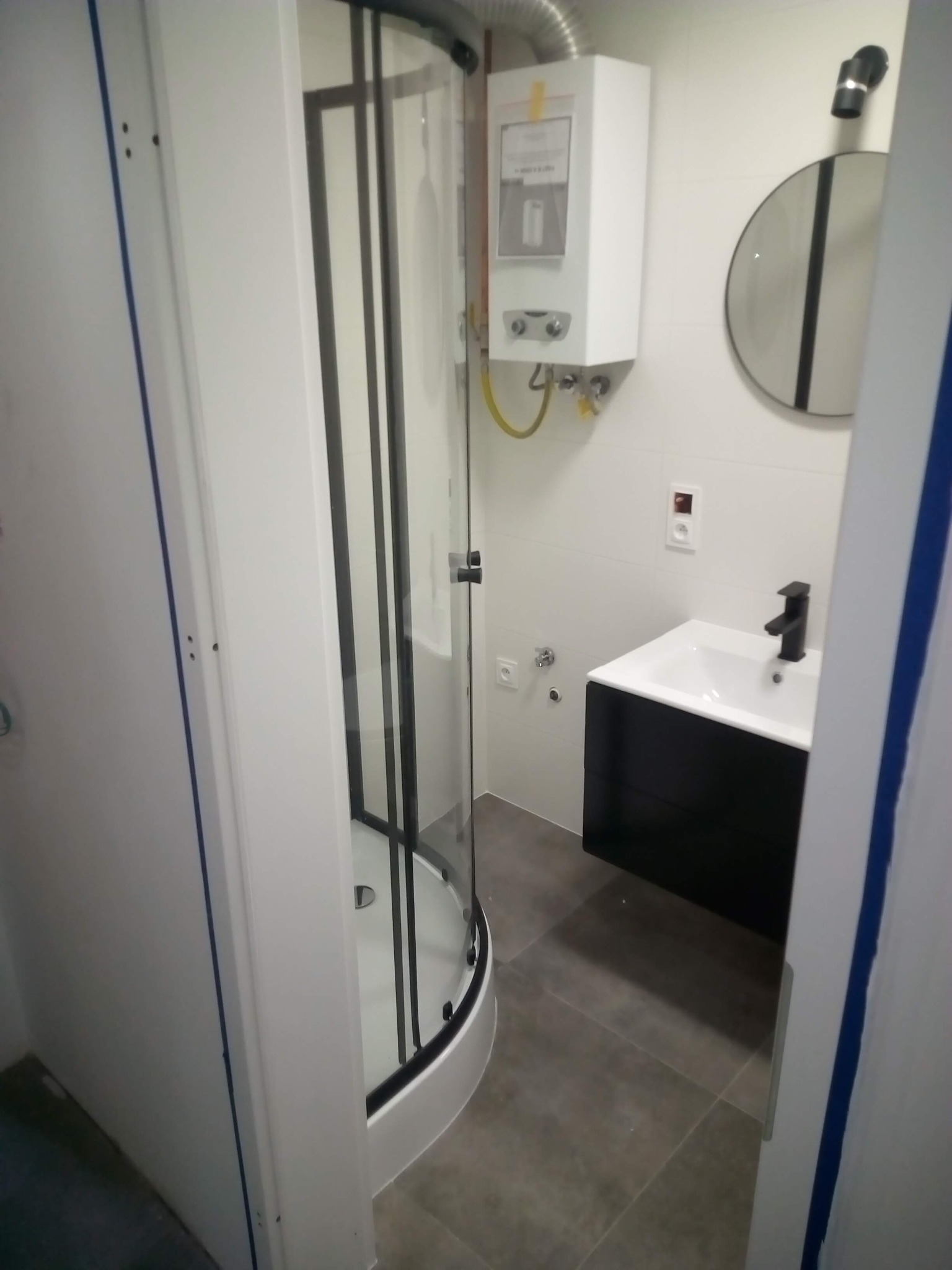 Nowoczesna łazienka po remoncie: kabina prysznicowa z czarnymi profilami, umywalka z szafką w kolorze czarnym, okrągłe lustro z czarnym paskiem, biały piecyk gazowy na ścianie z białymi płytkami.