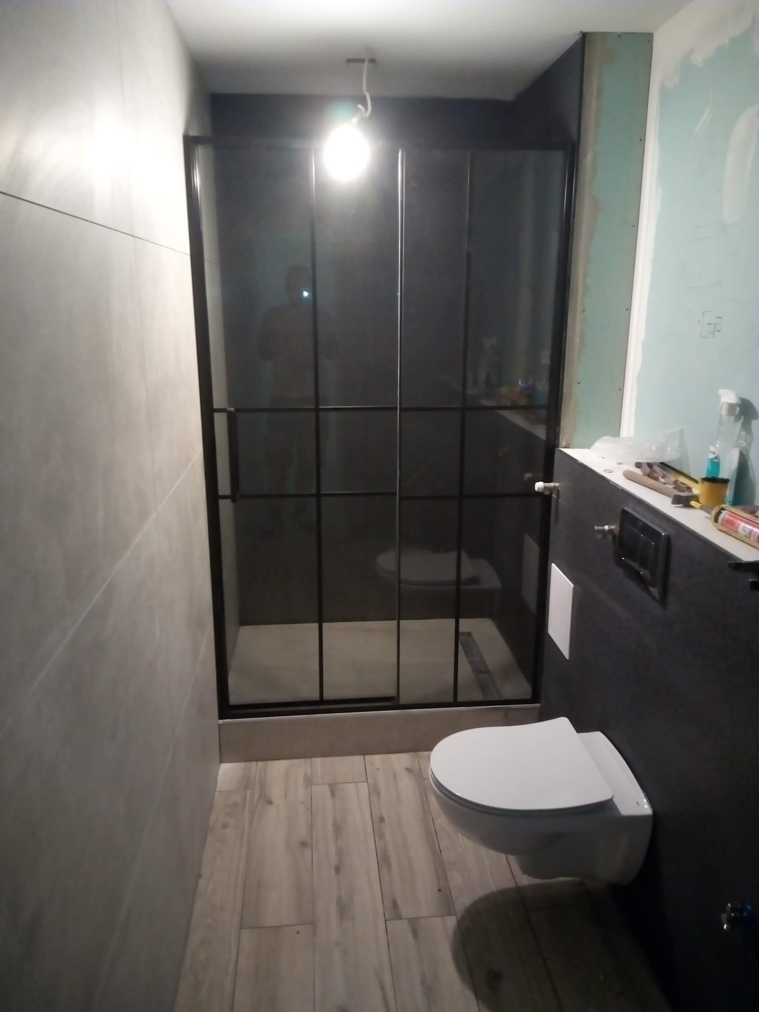 Niedokończona łazienka z płytkami w odcieniach szarości, kabiną prysznicową z czarnymi ramami i podwieszaną toaletą; widoczne narzędzia i materiały budowlane na blacie.