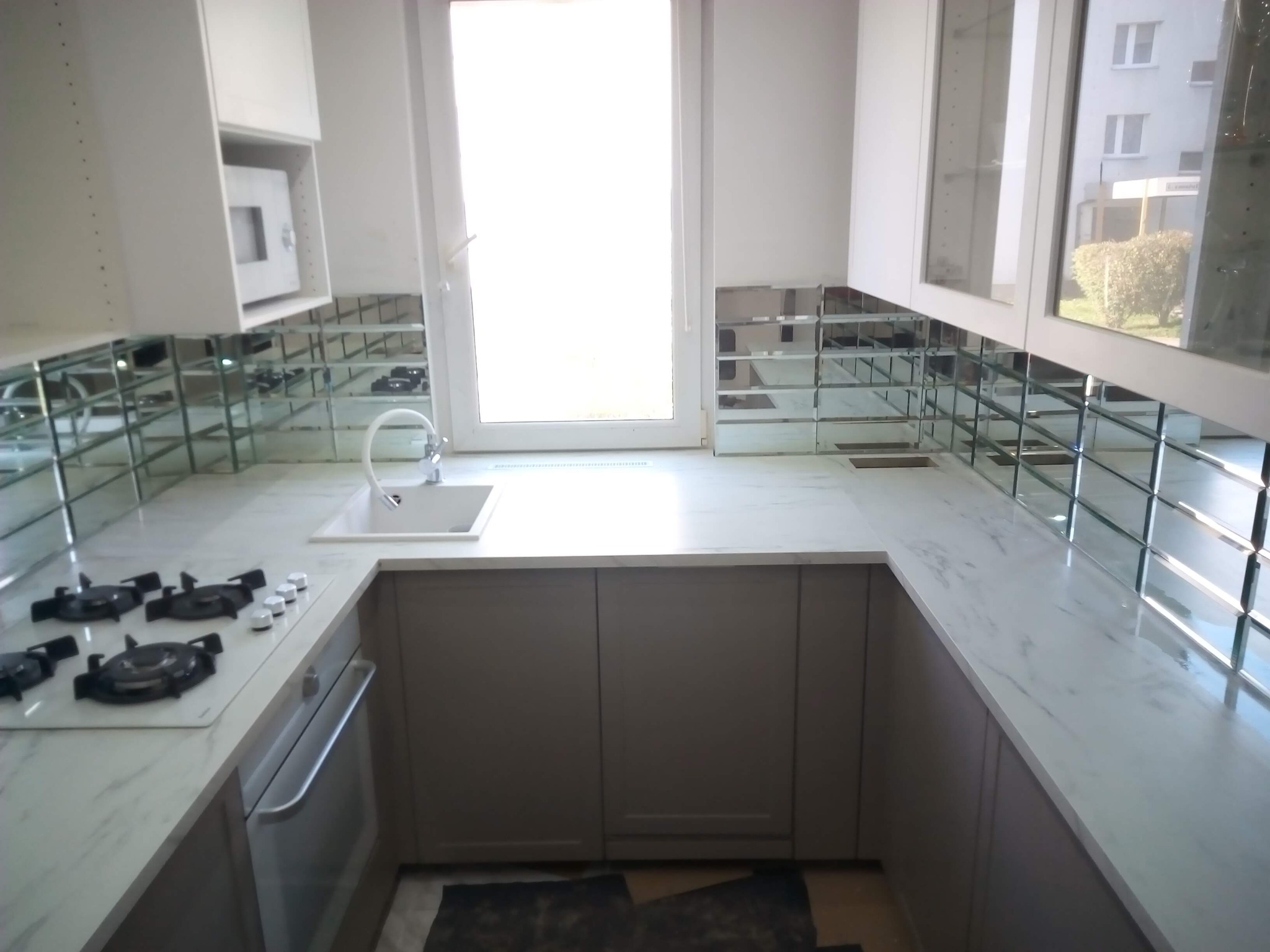 Nowoczesna kuchnia z białymi szafkami, marmurowym blatem i lustrzaną mozaiką na ścianie, widok na okno i zabudowany piekarnik.