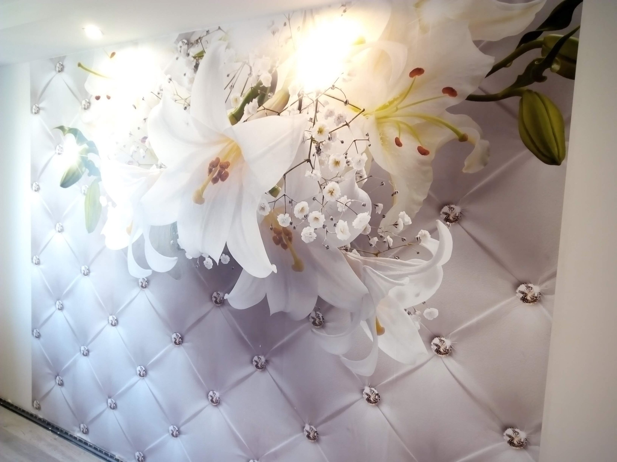 Fototapeta z motywem białych lilii i pikowanego wzoru z kryształkami, oświetlona od góry, tworząca elegancki efekt dekoracyjny na ścianie.