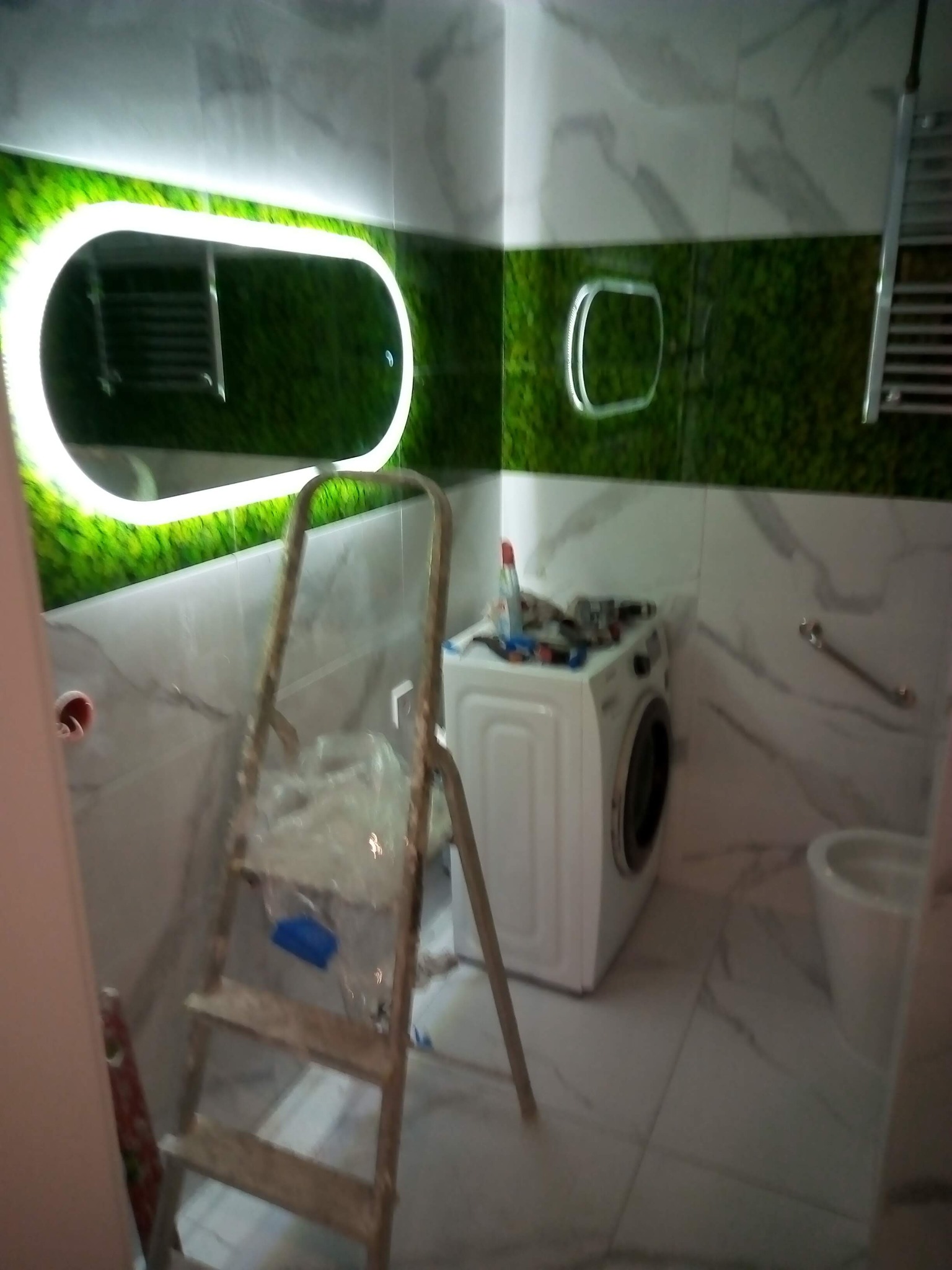 Łazienka w trakcie prac: drabina, pralka z narzędziami, podświetlane lustro owalne na tle zielonej ściany, białe płytki imitujące marmur.