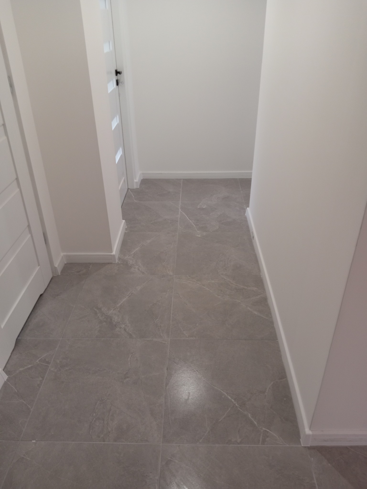 Szara, marmurkowa podłoga z płytek w korytarzu, białe ściany i listwy przypodłogowe, widoczne drzwi z białymi panelami.