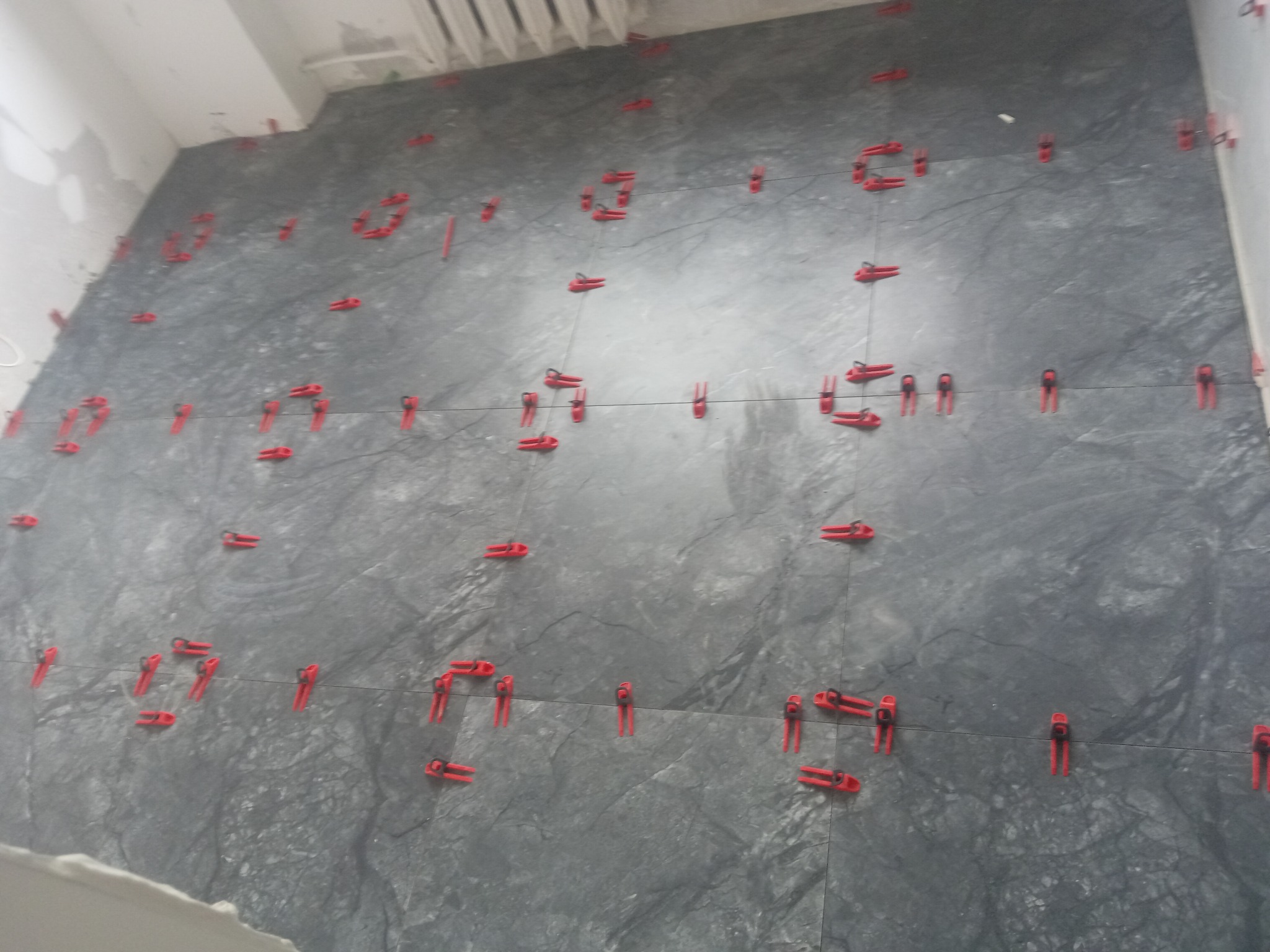 Świeżo położone, duże, szare płytki na ścianie, z czerwonymi krzyżykami dystansowymi, widoczne niedoskonałości fugi i surowy stan wykończenia góry ściany.