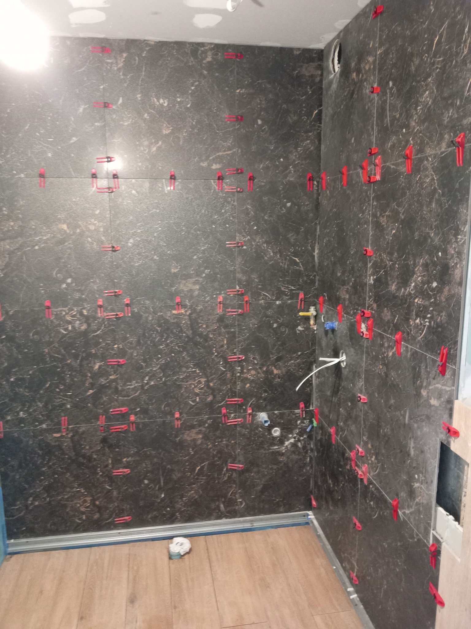 Ściany łazienki w trakcie układania płytek imitujących marmur, z użyciem czerwonych klipsów poziomujących, widoczne fragmenty instalacji wodnej i elektrycznej, podłoga wyłożona panelami...