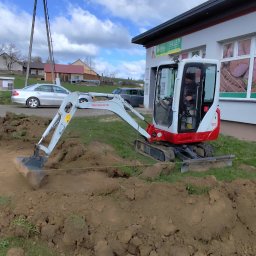 Damian Krówka - Mini koparka Takeuchi TB210 w trakcie wykopu ziemi obok budynku sklepu, widoczne narzędzia i wytyczone granice. Prace ziemne w słoneczny dzień.