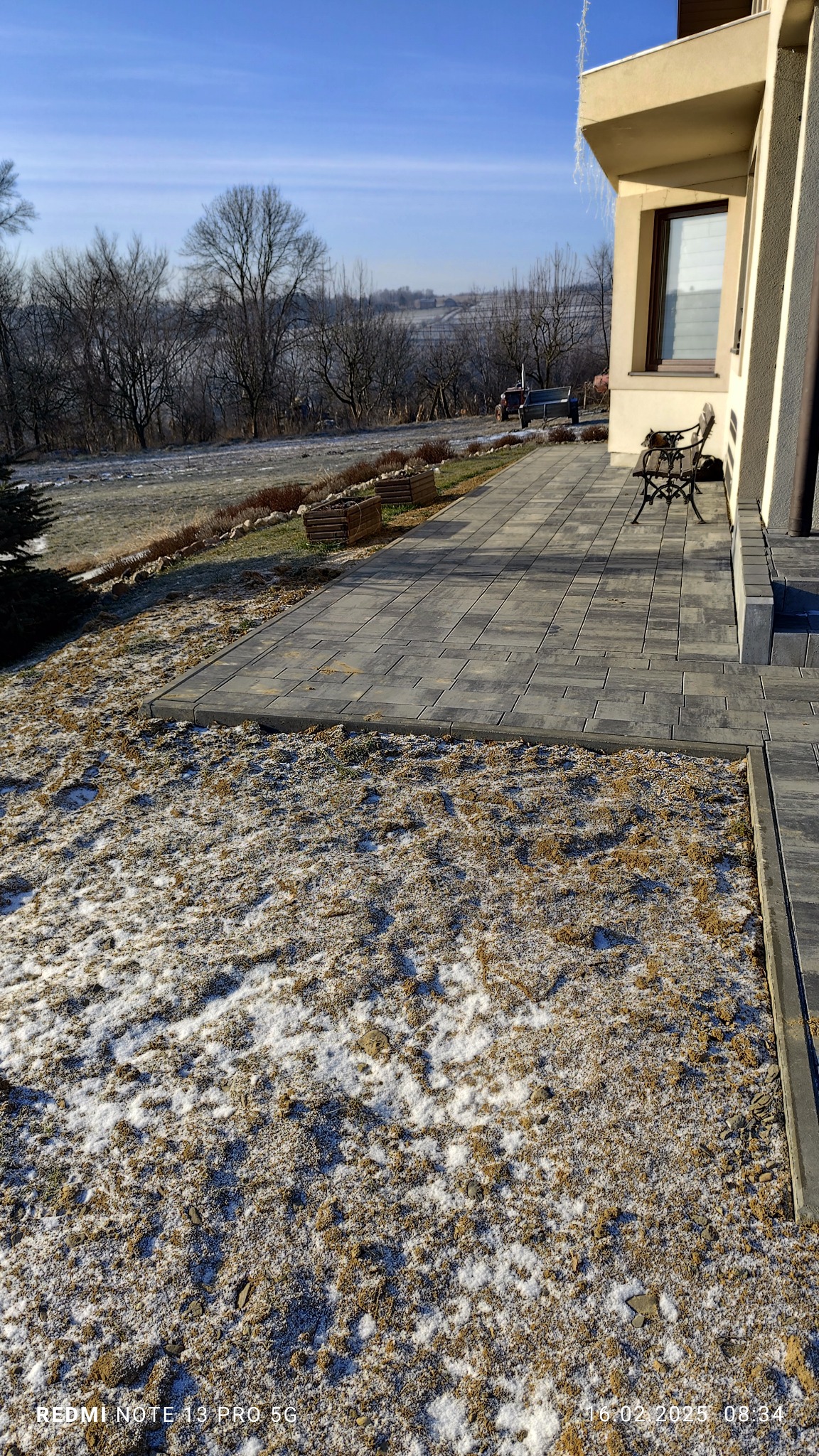 Ułożona szara kostka brukowa tworzy chodnik i taras przy domu w zimowej scenerii, z częściowo pokrytym śniegiem trawnikiem i widokiem na wzgórza w tle.