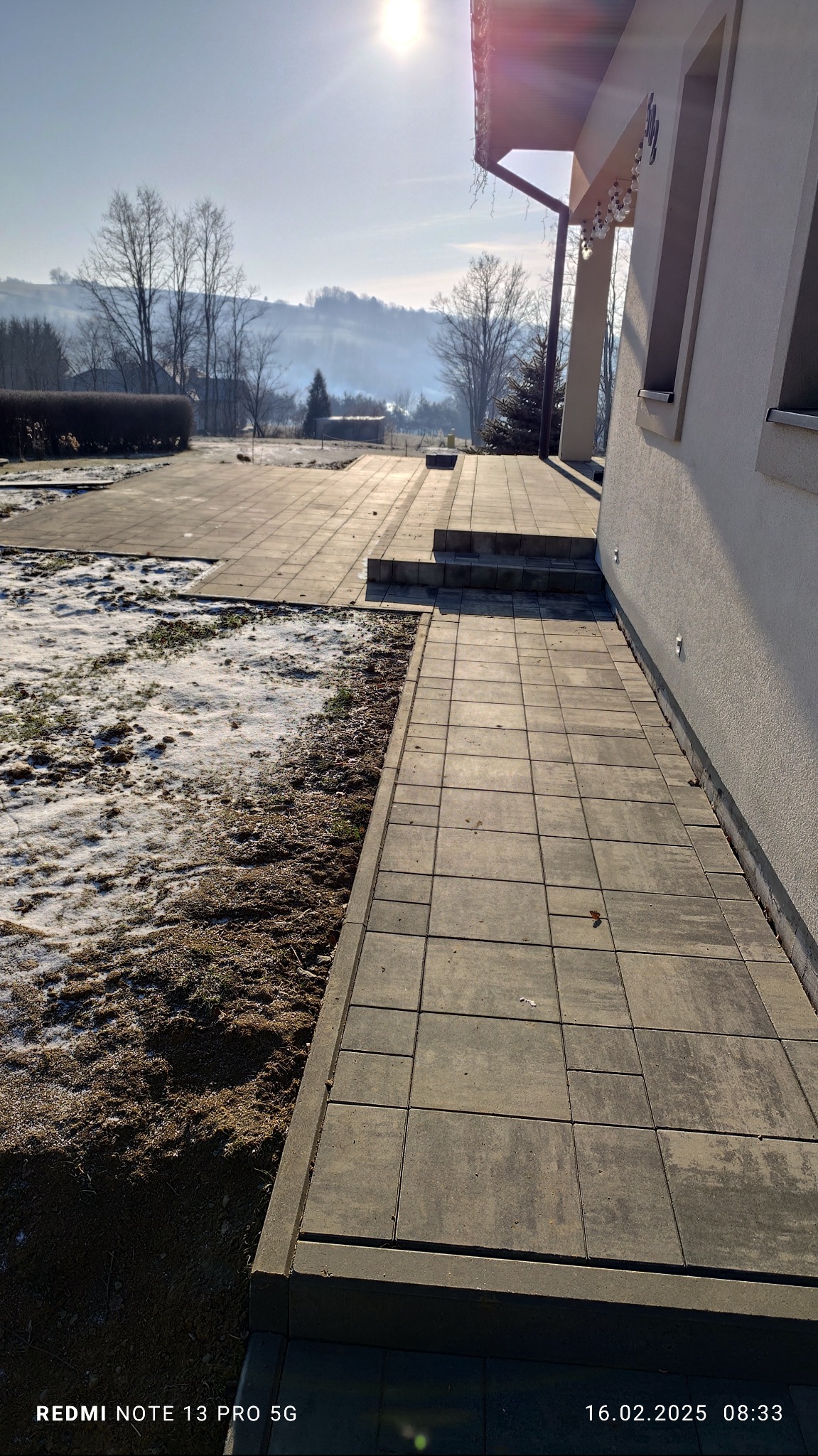 Ułożona kostka brukowa wzdłuż ściany domu z widocznymi schodami, częściowo pokryta śniegiem, z zalesionym wzgórzem w tle pod jasnym, zimowym słońcem.