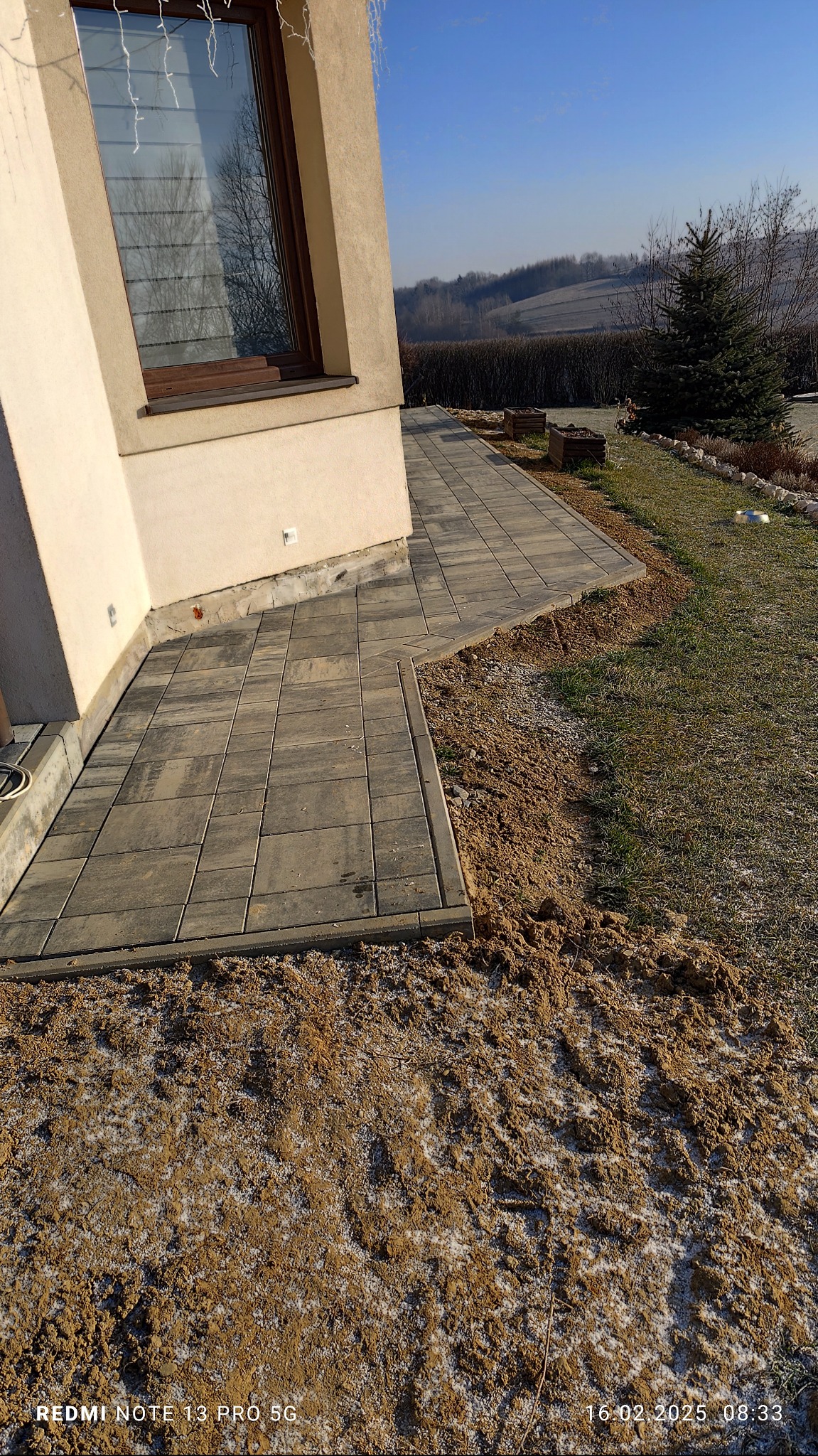 Ułożona szara kostka brukowa przy ścianie budynku z oknem, widoczny fragment niezagospodarowanego terenu z ziemią i trawą, w tle wzgórza i drzewa.