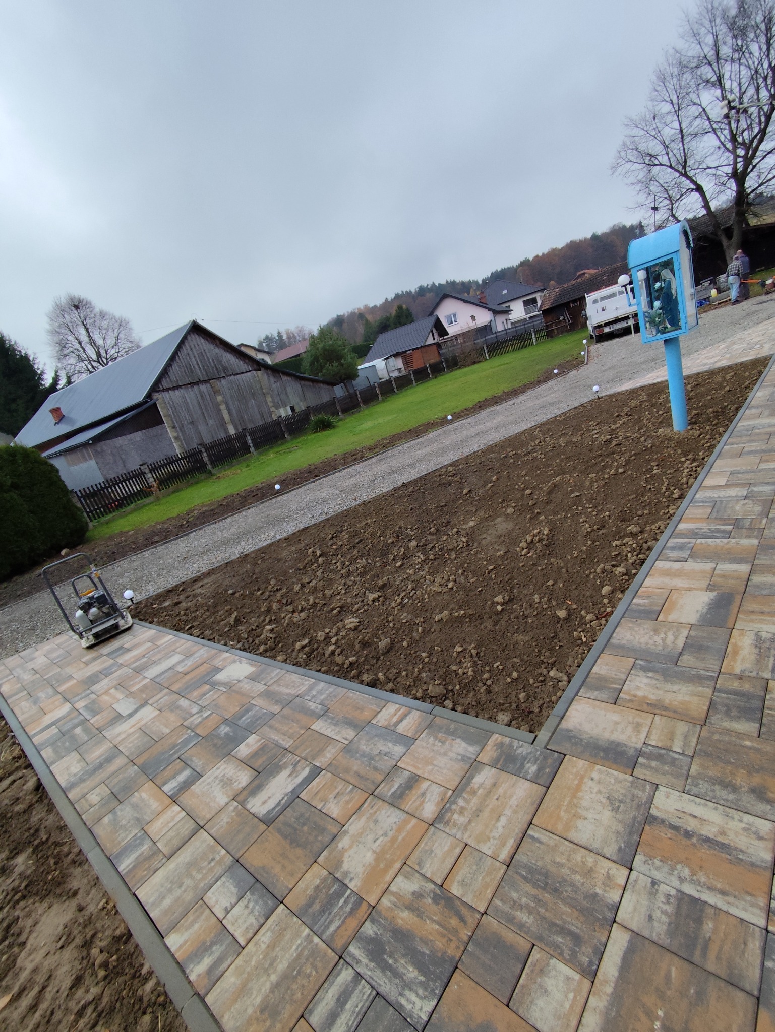 Ułożona kostka brukowa w odcieniach beżu i szarości, obok wyrównany teren pod trawnik. W tle budynek gospodarczy i droga z białym żwirem. Widoczna ubijarka.