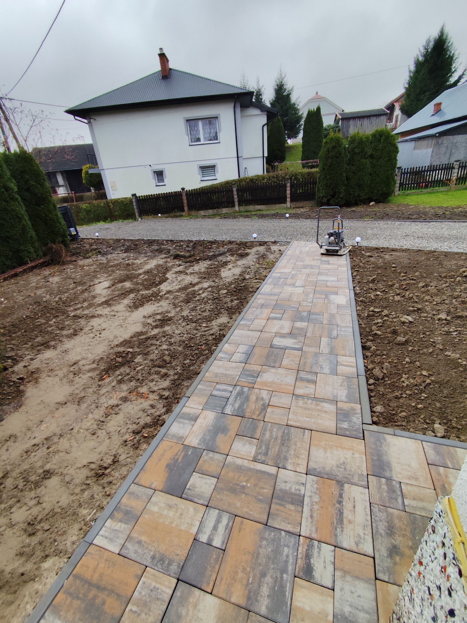 Ścieżka z kostki brukowej w odcieniach szarości i beżu, prowadząca przez ogród w kierunku domu. Widoczna ubijarka wibracyjna. Częściowo ułożona kostka.